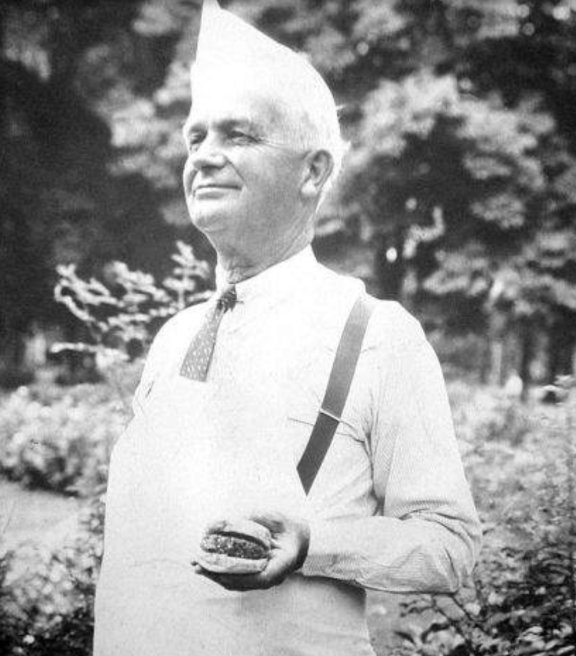 Older man in a light shirt with suspenders and a tall cone-shaped hat, standing outdoors and holding a hamburger.