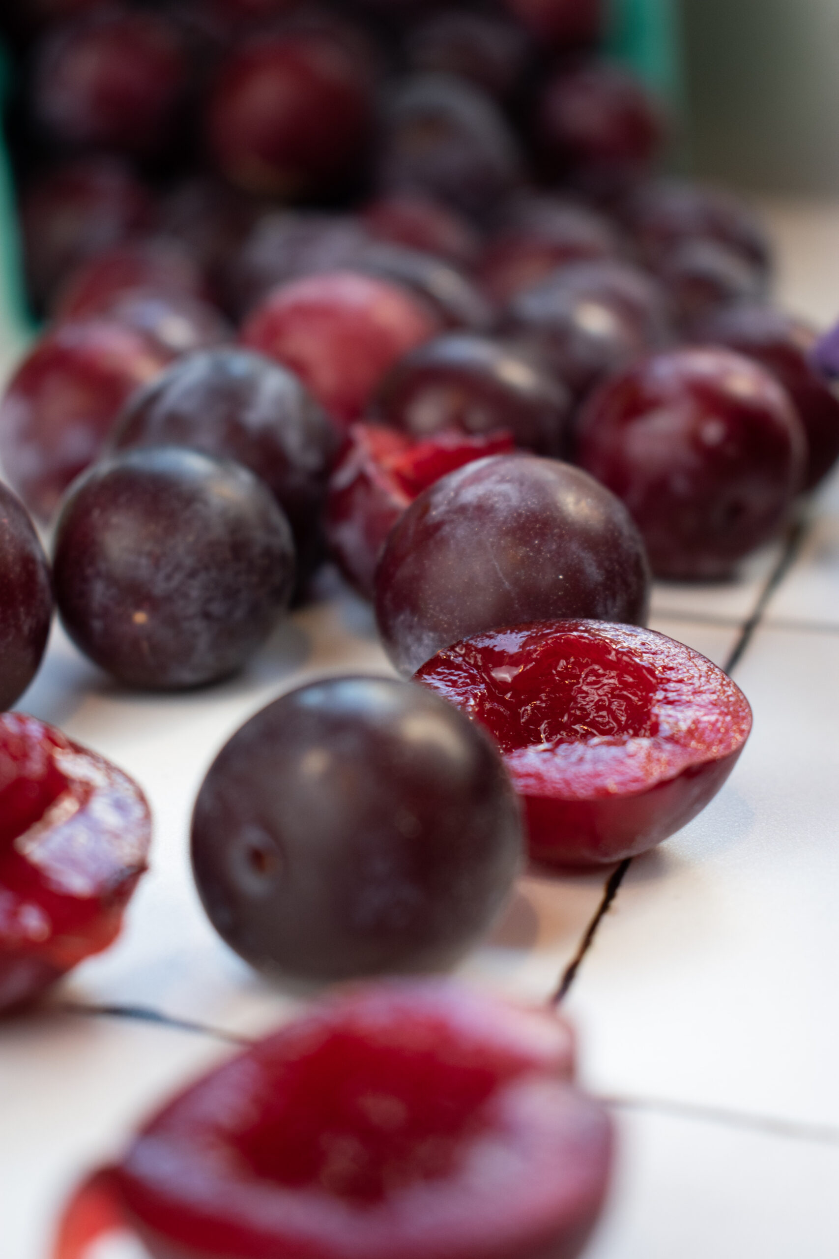 Close-up of glossy purple plums, some halved to show bright red flesh, arranged on a wire rack/tray.