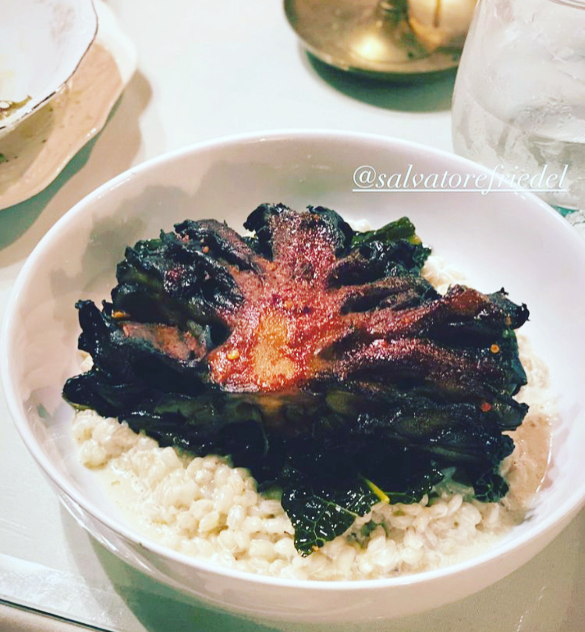 Creamy risotto in a white bowl topped with a flower-shaped mound of crispy, dark greens with a reddish center.